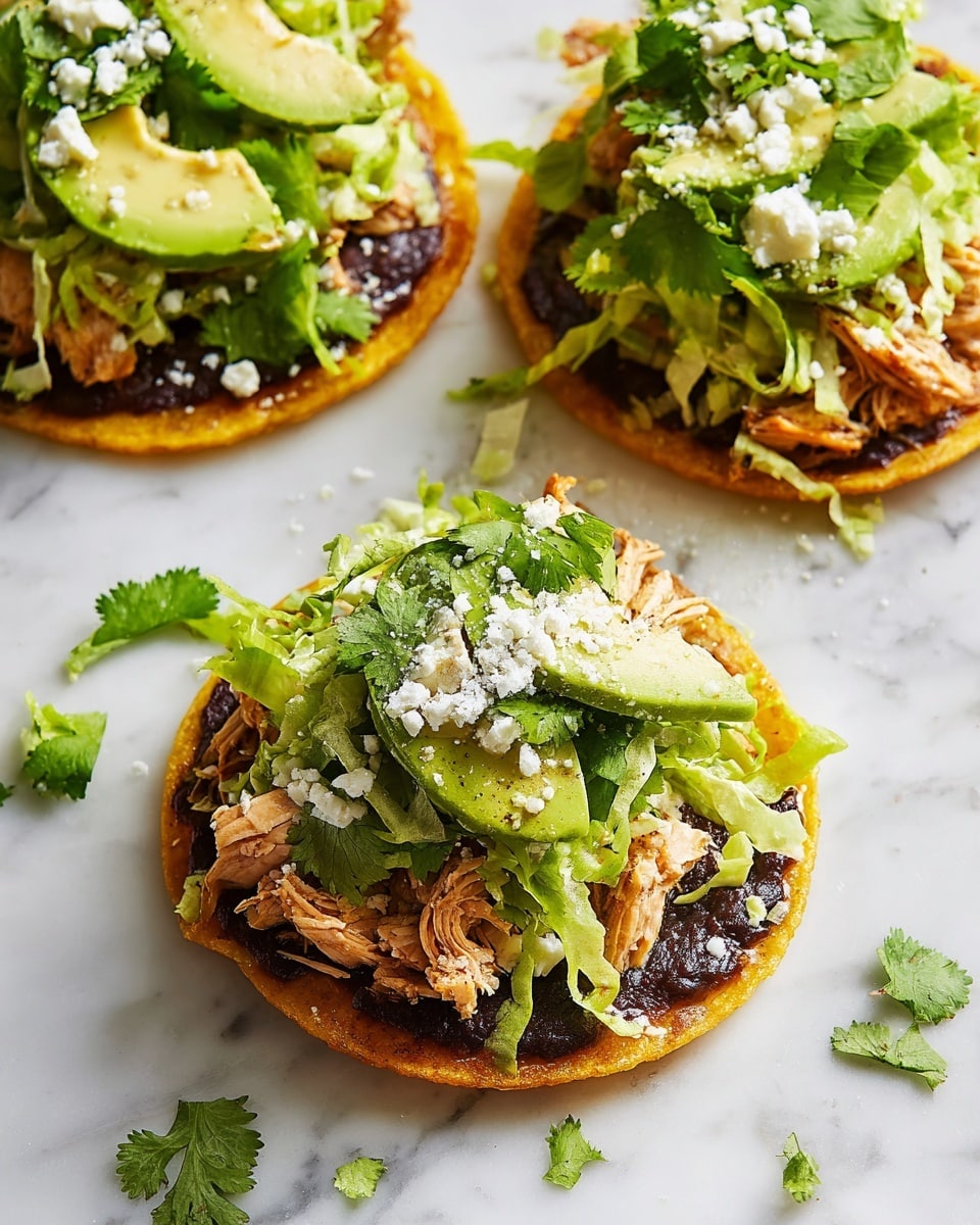 The image shows three small round tostadas on a white marbled surface. Each tostada has four layers: the bottom layer is a crispy golden yellow tortilla, topped with a dark, smooth black bean spread. Above this is a layer of shredded light brown chicken, then a generous topping of bright green shredded lettuce. On the top, there are a few slices of avocado, sprinkled with white crumbled cheese, and garnished with fresh green cilantro leaves. Small crumbs of cheese and pieces of lettuce are scattered around the tostadas. Photo taken with an iphone --ar 4:5 --v 7