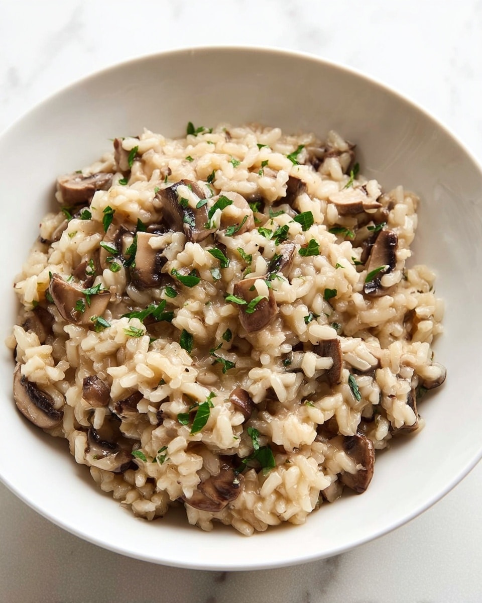 A close-up of a white bowl filled with creamy mushroom risotto, showing soft, light beige rice mixed with small chopped mushrooms in varying shades of brown and gray, with small pieces of green herbs scattered evenly on top. The risotto looks rich and moist, with the grains of rice clinging together in a slightly glossy texture. The bowl rests on a white marbled surface. photo taken with an iphone --ar 4:5 --v 7