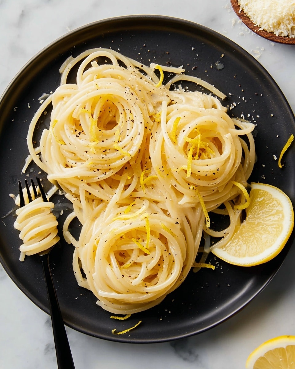 A black round plate holds three neatly twirled nests of creamy pale yellow spaghetti, each sprinkled lightly with ground black pepper and topped with a few thin, curly strips of bright yellow lemon zest. A wedge of fresh lemon with a vibrant yellow peel rests at the lower right side of the plate. A black fork sits near the bottom left side, with some spaghetti strands wrapped around its prongs. The plate is set on a white marbled surface with a small pile of grated cheese visible in the upper right corner. Photo taken with an iphone --ar 4:5 --v 7