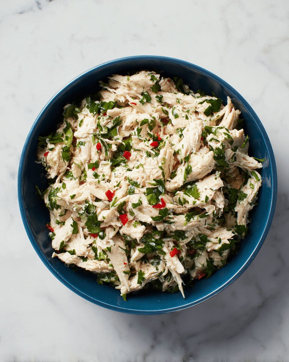 A close-up top view of a blue bowl filled with shredded cooked chicken mixed with small green leafy herbs and tiny red chili pieces; the chicken pieces are creamy white with a soft texture, while the herbs add fresh green flecks evenly spread throughout, and the red chili bits add small pops of bright red color; the bowl sits on a white marbled surface. photo taken with an iphone --ar 4:5 --v 7