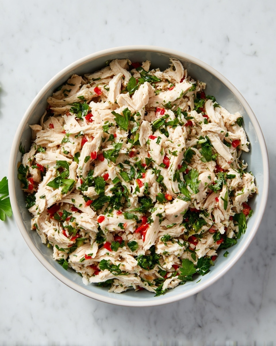 A close-up top view of a round white bowl filled with shredded white chicken mixed with chopped green herbs and small pieces of red pepper. The chicken is light beige with a moist texture, scattered evenly with fresh green herbs and tiny red bits, giving a colorful contrast. The bowl sits on a white marbled surface, creating a clean and bright background. photo taken with an iphone --ar 4:5 --v 7