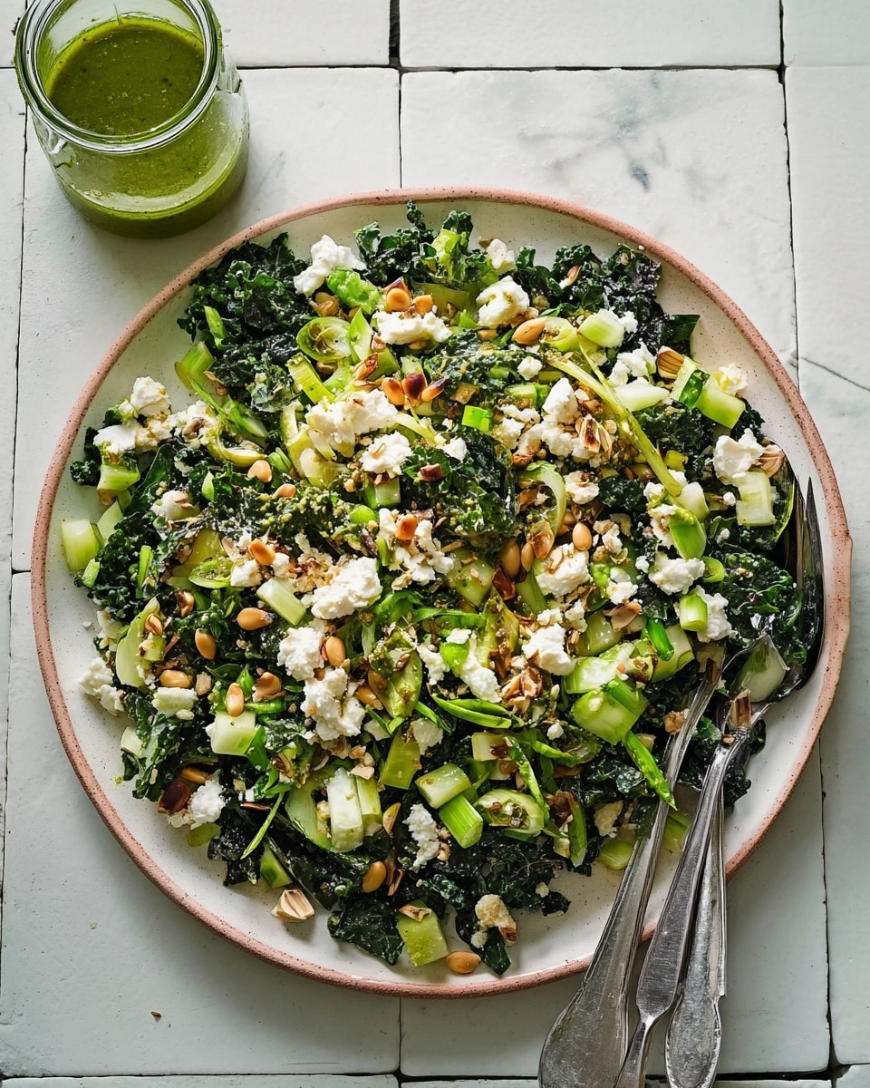 A large white plate filled with a fresh salad sits on a white marbled tiled surface. The salad has multiple layers, starting with dark green leafy kale at the bottom, mixed with light green chopped celery and cucumber pieces. On top, there are scattered dollops of soft white cheese and toasted light brown pine nuts. The salad is drizzled with a green herb dressing and garnished with slices of green onion. Two metal forks rest on the right side of the plate, partially on the salad. A small glass jar containing more green dressing is placed in the upper left corner. The overall look is fresh, vibrant, and colorful. Photo taken with an iphone --ar 4:5 --v 7