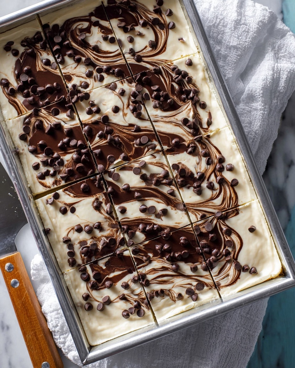 A rectangular metal pan holds a dessert with two visible layers: a smooth, creamy pale white layer on the bottom and a swirled dark brown chocolate layer spread thinly on top in irregular patterns. The top is generously sprinkled with small, round chocolate chips scattered evenly across the surface. The dessert is cut into neat square slices, and a large metal spatula with a wooden handle rests beside the pan on a white marbled surface. A white cloth is partially placed under the pan. Photo taken with an iphone --ar 4:5 --v 7