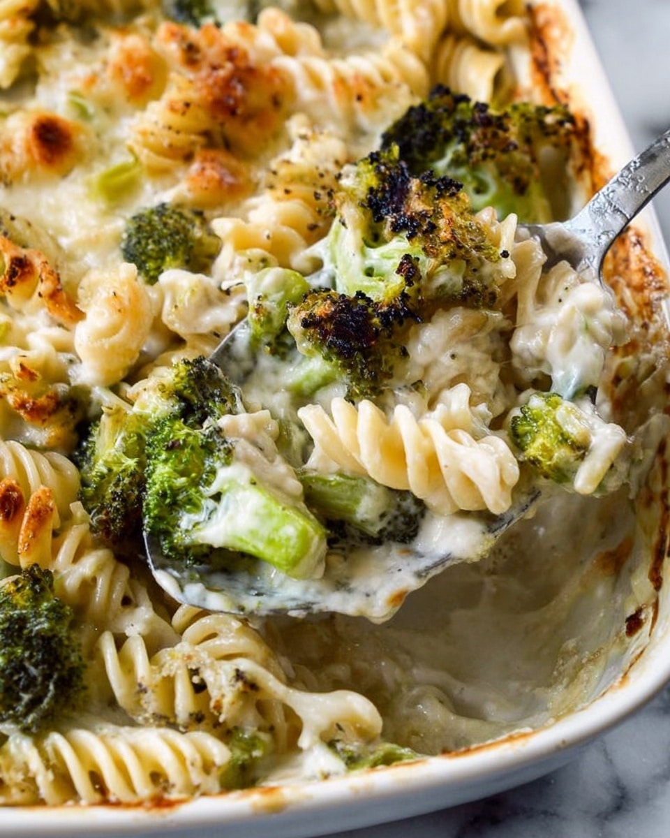 The image shows a close-up of a pasta dish with rotini noodles mixed with green broccoli pieces, all covered in a creamy white cheese sauce. The top layer is slightly browned and bubbly, showing melted cheese with some crisp spots. The pasta looks soft and the broccoli is tender with some darker, roasted edges. A metal spoon scoops up a mix of noodles and broccoli from the dish, resting on a white marbled surface. photo taken with an iphone --ar 4:5 --v 7