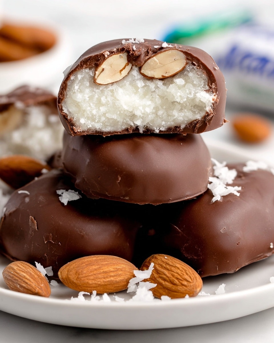 The image shows a close-up of chocolate-covered almond candies stacked on a smooth white plate against a white marbled background. Each candy has three visible layers: a shiny, medium brown chocolate coating on the outside, a white, slightly crumbly and fluffy center, and whole almonds embedded near the top inside the chocolate layer, visible where the candy is cut in half. The almonds have a natural light brown skin and a smooth inner texture. Around the candies, there are a few whole almonds and small pieces of white coconut flakes scattered on the plate. Photo taken with an iphone --ar 4:5 --v 7