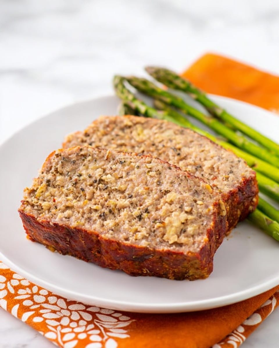 Two thick slices of meatloaf with a golden brown crust are placed in the center of a white plate. The inside of the meatloaf shows a textured mix of ground meat and oats, with small specks of seasoning visible throughout. Behind the meatloaf, several vibrant green asparagus spears rest diagonally across the plate, adding a fresh contrast in color. The plate sits on an orange napkin with a white floral pattern, all set on a white marbled surface. photo taken with an iphone --ar 4:5 --v 7