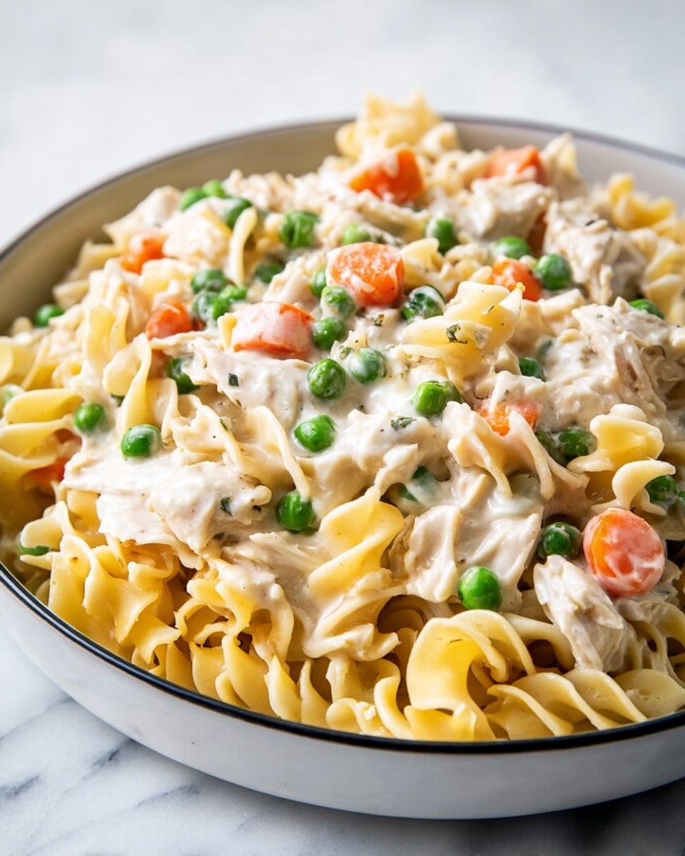 Close-up of creamy pasta dish in a white bowl on a white marbled surface, showing three layers: the bottom layer has yellow egg noodles with ruffled edges, the middle layer features chunks of white chicken mixed with the noodles, and the top layer includes small bright green peas and diced orange carrots all covered in a smooth, pale cream sauce that coats the pasta and vegetables evenly, creating a rich and inviting texture, photo taken with an iphone --ar 4:5 --v 7