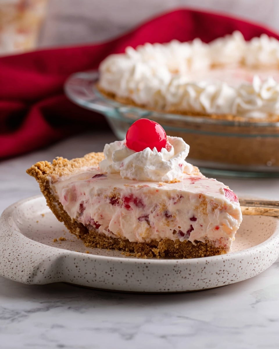 The image shows a slice of creamy pie on a white speckled dish with a handle. The pie has three visible layers: a thick, light brown crumbly crust at the bottom, a thick creamy filling with pink and red pieces mixed inside, and a white whipped cream topping with a single bright red cherry placed on top. In the background, there is a round clear glass pie dish with the rest of the pie, topped with whipped cream. A red cloth is slightly visible on the left side, and the setting is on a white marbled surface. Photo taken with an iphone --ar 4:5 --v 7