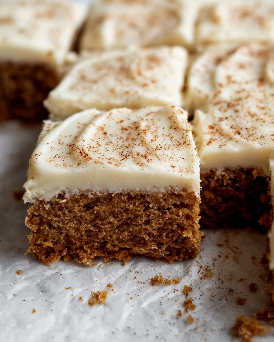 The image shows close-up square pieces of a moist, brown cake with a thick layer of creamy white frosting spread evenly on top. The frosting has a smooth texture with slight swirls and is sprinkled lightly with cinnamon powder. The cake has a soft, slightly crumbly texture and sits on a white marbled surface with some crumbs scattered around. Photo taken with an iphone --ar 4:5 --v 7