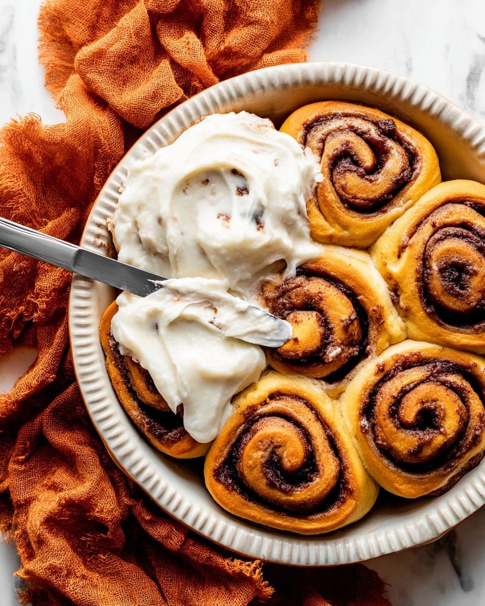 The image shows a close-up of seven golden-brown cinnamon rolls arranged tightly in a white ceramic round baking dish with a ridged edge. The rolls have visible layers swirling with dark brown cinnamon filling, and three of the rolls on the left side are partially covered with thick, creamy white frosting that has small specks, giving it a smooth and rich texture. A silver knife with some frosting on it rests on the right side of the dish, spreading the frosting onto one of the rolls. The baking dish is placed on a white marbled surface with a crumpled burnt orange cloth underneath it. photo taken with an iphone --ar 4:5 --v 7