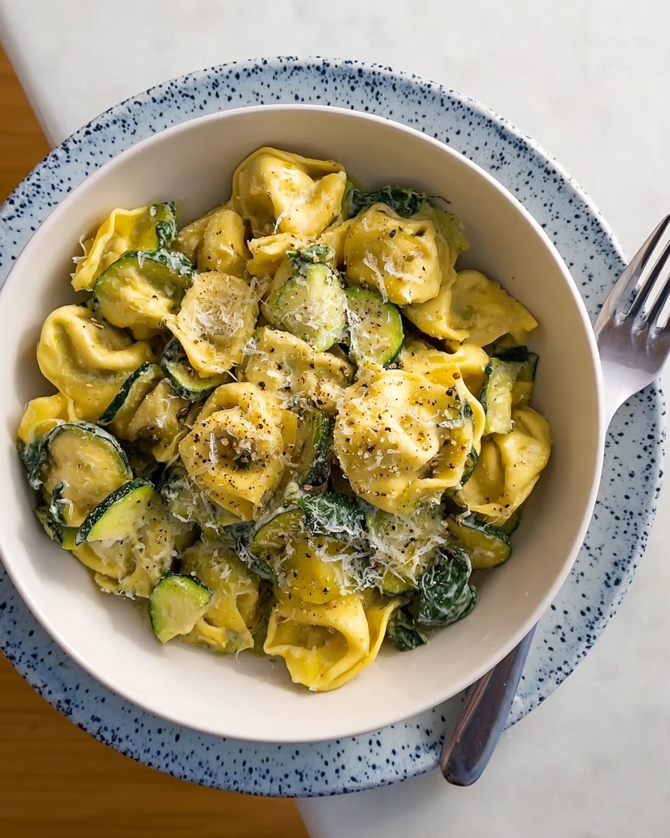 A white bowl filled with tortellini pasta that has a mix of yellow and green colors, showing a soft and slightly slightly wrinkled texture, and includes cooked spinach leaves and thin sliced green zucchini mixed throughout. The pasta is topped with a sprinkle of grated white cheese and a light dusting of black pepper, giving a speckled look. The bowl rests on a larger white plate with blue speckles, and a silver fork lies beside it, all set on a smooth white marbled surface. photo taken with an iphone --ar 4:5 --v 7