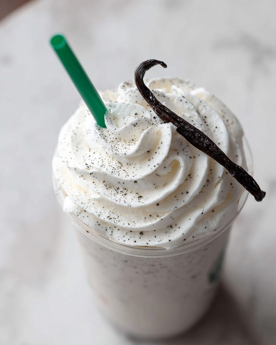 A creamy drink fills a clear cup, topped with a thick, fluffy layer of white whipped cream that has small black specks scattered across it. On top of the whipped cream sits a whole dark brown vanilla bean pod, curved elegantly. A green straw sticks out from the whipped cream, contrasting with the light colors below. The cup is placed on a white marbled surface. photo taken with an iphone --ar 4:5 --v 7