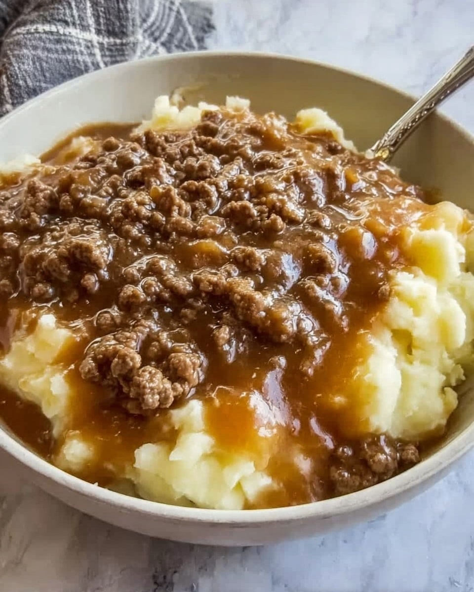 The image shows a close-up of a bowl filled with mashed potatoes topped with ground beef mixed into brown gravy. The mashed potatoes form the bottom layer with a soft, creamy, and slightly lumpy texture in a light yellow color. On top is a thick layer of cooked ground beef mixed with rich brown gravy that looks glossy and smooth, covering most of the mashed potatoes. The bowl is white and placed on a white marbled surface with a spoon resting inside the bowl, visible at the top edge. photo taken with an iphone --ar 4:5 --v 7