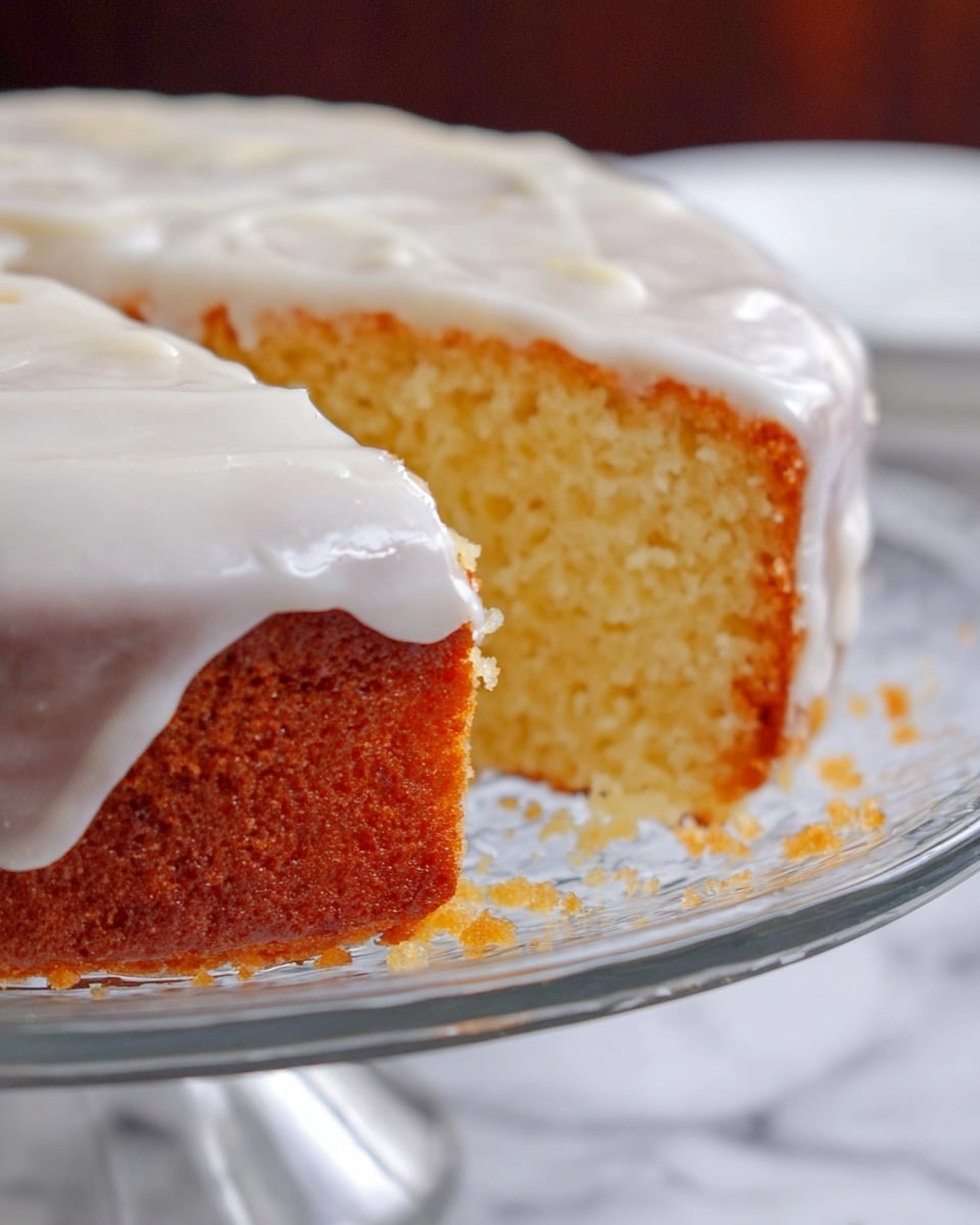 A close-up view of a single-layer round cake with a golden-brown crust and a moist, light yellow inside. The cake is topped with a thick layer of smooth, white icing that slightly drips over the edges. It is placed on a clear glass cake stand, which sits on a surface with a white marbled texture. The cut side shows crumbs scattered lightly around the base of the cake. photo taken with an iphone --ar 4:5 --v 7