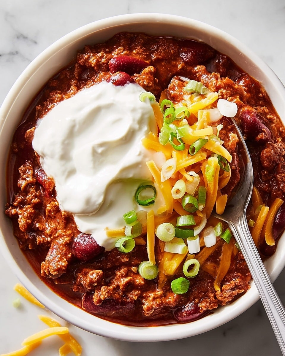 A white bowl filled with rich, thick chili showing two layers: a deep red-brown mix of ground meat and kidney beans at the bottom, topped with bright orange shredded cheddar cheese and small pieces of chopped green onions and white onions scattered on top. On one side, smooth white sour cream is being scooped up with a silver spoon, creating a creamy contrast against the chili. The white marbled surface underneath highlights the colors of the dish, with some small bits of cheese and onion on the surface for added detail. photo taken with an iphone --ar 4:5 --v 7