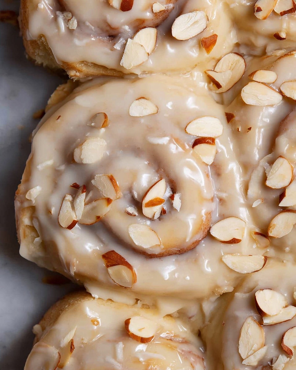 The image shows multiple soft cinnamon rolls covered in a thick layer of creamy off-white icing. Scattered on top are many thin almond slices with light brown edges, adding texture and contrast to the smooth icing. Each roll is visible with their swirled dough pattern beneath the icing, showing a pale golden brown color. The rolls are closely packed, and the background is a white marbled texture. photo taken with an iphone --ar 4:5 --v 7