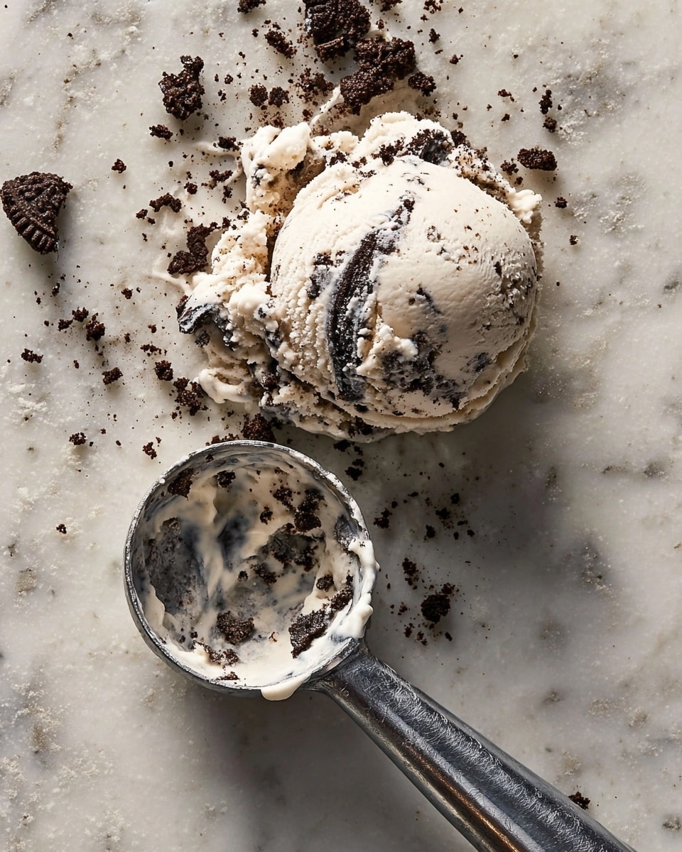 A single scoop of cookies and cream ice cream with visible chunks of dark chocolate cookie pieces in creamy white ice cream sits on a white marbled surface, slightly melting with some cookie crumbs scattered around it. Next to the scoop is a metal ice cream scoop with remnants of the ice cream inside, showing a mix of creamy white and dark cookie bits, the scoop handle extends diagonally towards the lower right of the image. Photo taken with an iphone --ar 4:5 --v 7