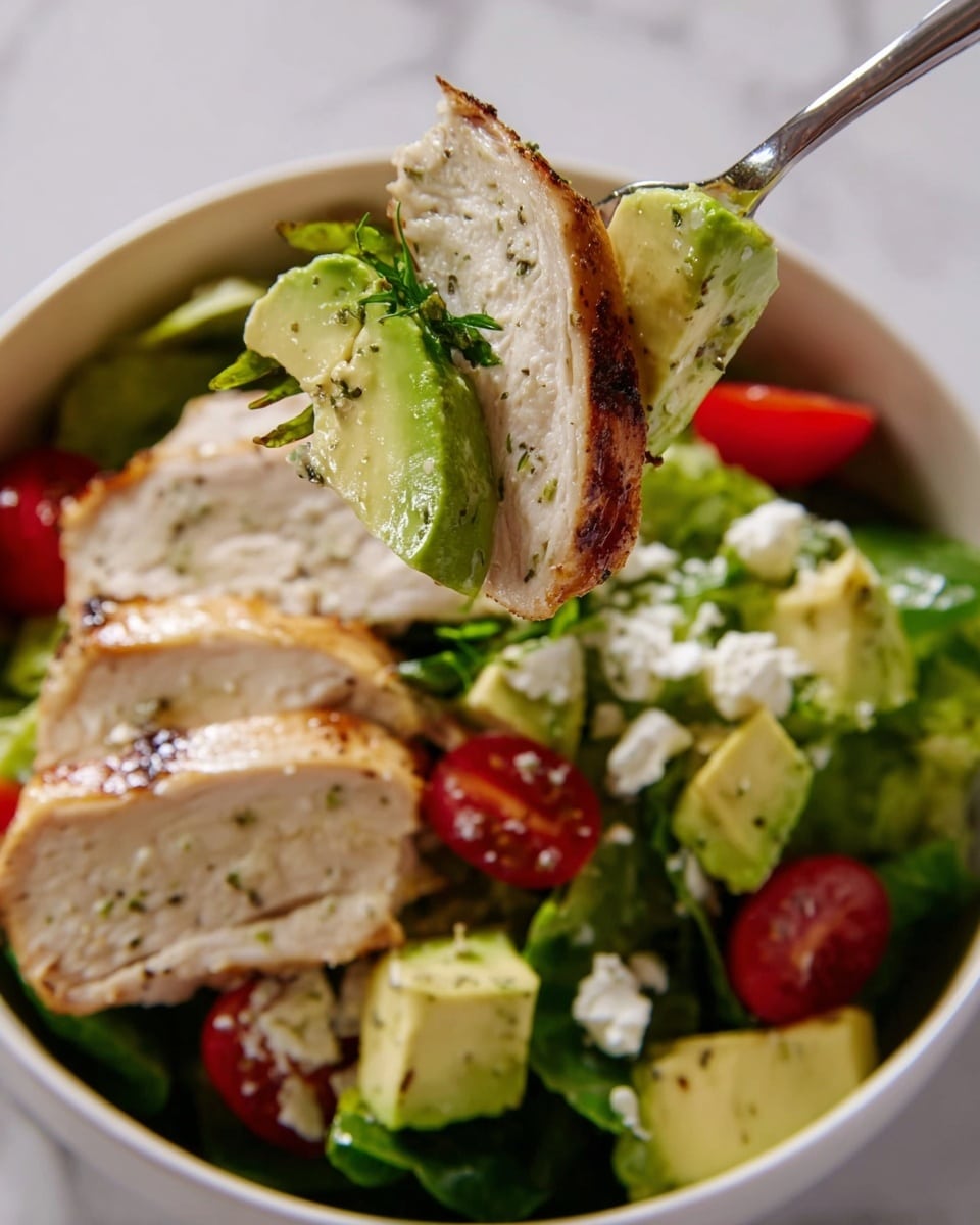 A white bowl filled with a fresh salad showing layers of green lettuce and spinach leaves at the bottom, bright red cherry tomato halves scattered among the greens, and creamy light green avocado chunks sprinkled with small pieces of white cheese. On top, thick slices of grilled chicken breast with a golden-brown sear and light seasoning are neatly arranged. A silver fork holds a bite-sized portion of the salad with a piece of grilled chicken, avocado, cherry tomato, and leafy greens lifted above the bowl. The scene is set on a white marbled texture. photo taken with an iphone --ar 4:5 --v 7