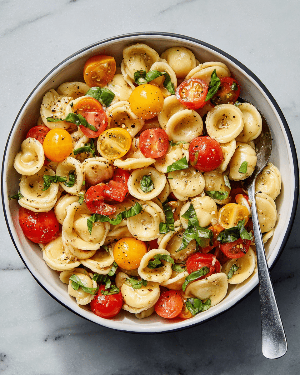 In a white bowl, there is a pasta dish made of light beige round orecchiette pasta mixed with halved red and yellow cherry tomatoes, and green basil leaves scattered throughout. The pasta and vegetables have a light coating of olive oil, sprinkled with black pepper and small bits of garlic, giving the dish a glossy and fresh look. A silver spoon is resting inside the bowl on the right side. The bowl is placed on a white marbled textured surface. photo taken with an iphone --ar 4:5 --v 7