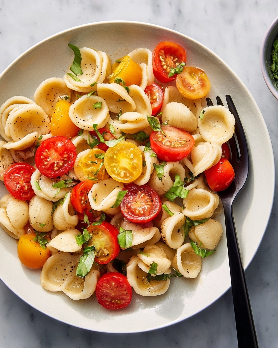 A white plate holds a serving of orecchiette pasta mixed with halved red and yellow cherry tomatoes. The pasta pieces are pale beige with a slightly glossy texture, scattered evenly throughout the dish. Small green basil leaves are sprinkled on top, adding a fresh touch. A black fork rests on the edge of the plate, with a few pasta pieces and a halved tomato speared on it. The plate sits on a white marbled surface. photo taken with an iphone --ar 4:5 --v 7
