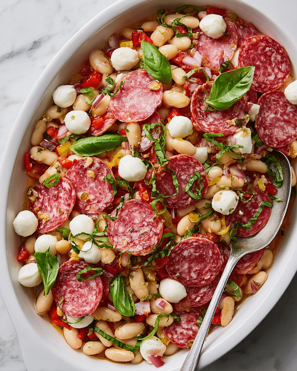 A white oval dish shows a colorful mixture of ingredients layered together. The base layer has white beans with a soft, creamy texture, topped with thin, round slices of pinkish-red salami with white fat specks. Scattered around are small, smooth, white mozzarella balls, mixed with small pieces of red and yellow pepper and diced red onion. Fresh, thinly sliced green basil leaves are spread evenly across the top, adding color contrast. A silver spoon rests on the right side inside the dish, partially buried under the salad, all set against a white marbled surface. Photo taken with an iphone --ar 4:5 --v 7
