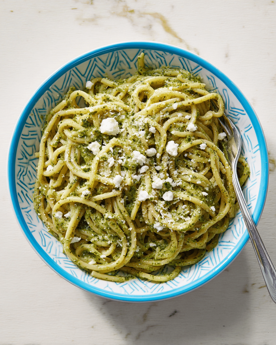 A white bowl with a blue geometric pattern around the edge holds a serving of thick, creamy green pesto pasta. The pasta strands are twisted and coated evenly in a light green, slightly oily pesto sauce with visible flecks of herbs. Small crumbles of white cheese are scattered on top, adding texture and contrast. A silver fork rests inside the bowl on the right side. The bowl is placed on a white marbled surface. photo taken with an iphone --ar 4:5 --v 7