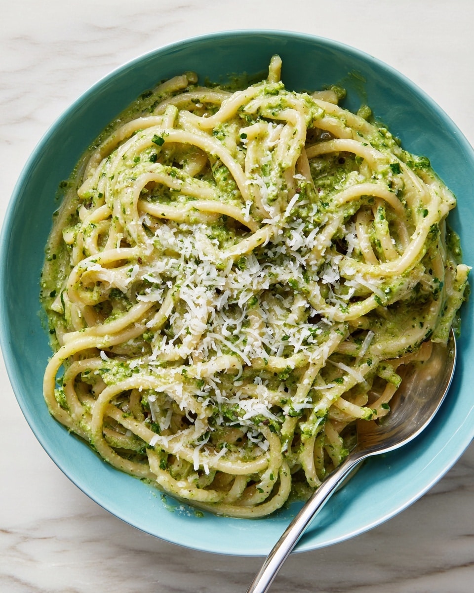 This image shows a close-up of pasta coated in a smooth, light green sauce with bits of finely chopped herbs. The pasta noodles are long, thick, and slightly twisted, with the sauce evenly covering each strand. White grated cheese is sprinkled liberally on top, adding a slightly crumbly texture that contrasts with the creamy sauce. A silver spoon is partially visible on the lower right corner, resting in the pasta. The dish is set against a white marbled background. photo taken with an iphone --ar 4:5 --v 7