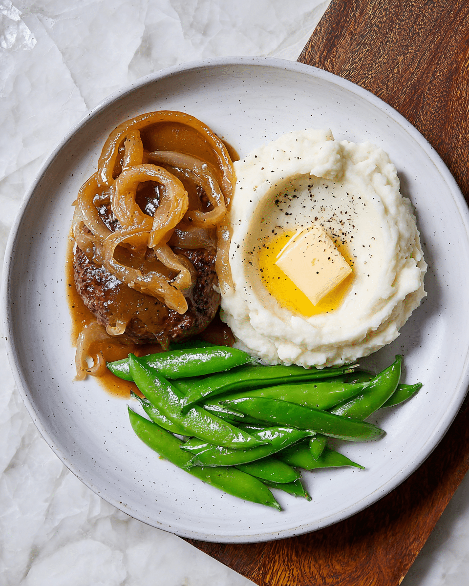 The dish shows one scoop of creamy white mashed potatoes molded in a smooth swirl with a small square of melting yellow butter in the center, lightly sprinkled with black pepper. To the left is a browned, cooked burger patty covered with a thick, glossy brown onion gravy topped with translucent cooked onion rings. On the right side of the plate, there is a cluster of shiny, bright green sugar snap peas that look fresh and crisp. All these ingredients rest on a large white plate with a faint texture visible, placed on a white marbled surface. photo taken with an iphone --ar 4:5 --v 7