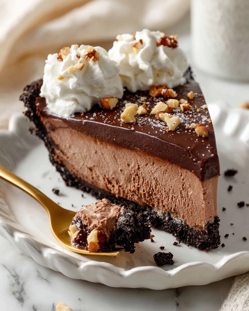 The image shows a slice of chocolate mousse pie on a white speckled plate with a ruffled edge, set on a white marbled surface. The pie has three layers: a dark chocolate crumb crust at the bottom, a thick creamy chocolate mousse filling in the middle, and a glossy, darker chocolate ganache topping sprinkled with chopped nuts and flaky sea salt. On top of the slice are two swirls of white whipped cream. A bite of the pie, with the mousse and crust visible, is placed on a gold fork beside the slice. Photo taken with an iphone --ar 4:5 --v 7