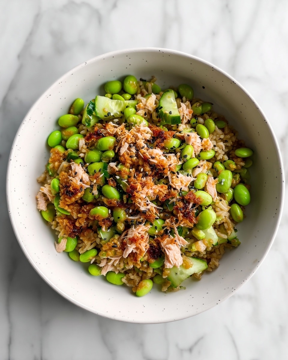 A white bowl filled with a colorful mix of food sits on a white marbled surface. The bottom layer is light brown rice with a slightly crispy texture visible. Scattered throughout are bright green edamame beans and thin slices of cucumber, adding fresh green touches. On top, there are flakes of light pink tuna with some browned, caramelized bits mixed in, and a sprinkling of small black sesame seeds. The food looks well mixed but you can still see the different textures and colors clearly. photo taken with an iphone --ar 4:5 --v 7