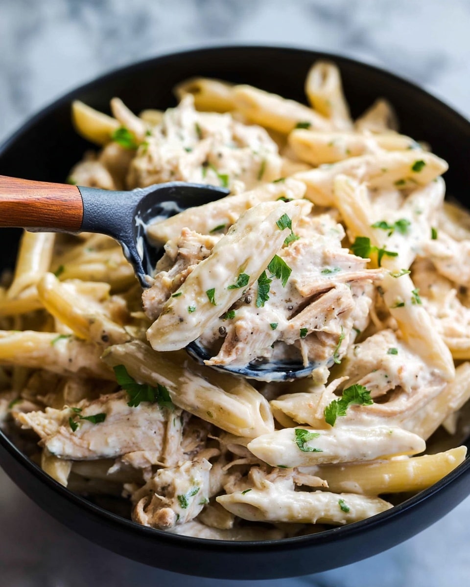 A close-up view of creamy penne pasta coated in a thick white sauce with small green herb pieces scattered on top. The pasta is mixed with shredded light brown chicken pieces, all sitting inside a black bowl. A black spoon with a wooden handle scoops up a portion, showing the smooth texture of the sauce clinging to the pasta and chicken. The background has a white marbled texture. photo taken with an iphone --ar 4:5 --v 7