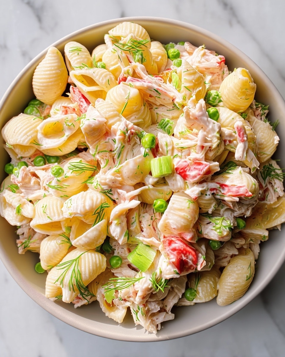 A close-up view of a pasta salad served in a white bowl, featuring three main layers: the bottom layer is made up of shell-shaped pasta in a light yellow color with a smooth texture, the middle layer consists of shredded white chicken mixed with creamy dressing that coats the pasta, and the top layer is garnished with bright green peas, chopped celery pieces, small sprigs of fresh dill, and bits of red bell pepper, adding pops of color and freshness. The overall look is creamy and inviting with light contrasts between the pale pasta and vibrant vegetables, all set on a white marbled surface. photo taken with an iphone --ar 4:5 --v 7