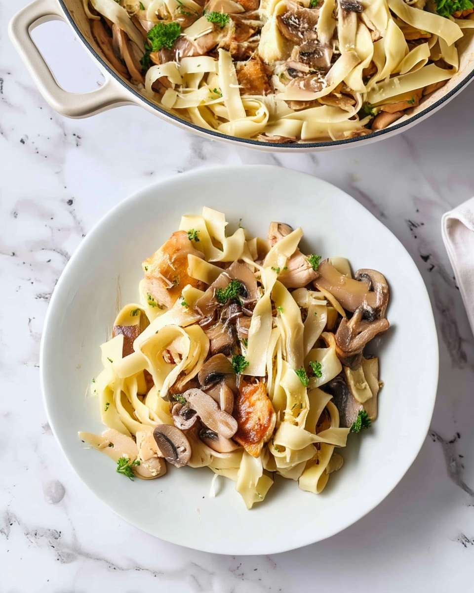 A white plate holds a portion of pasta with three main layers: the bottom layer is long, flat, cream-colored noodles; the middle layer is light brown cooked mushrooms cut into slices; scattered among these are pieces of golden-brown cooked chicken, adding texture and contrast; the top layer is a sprinkling of fresh green parsley and small shreds of white cheese. Above and behind the plate, a white pan with a single handle contains more of the same pasta dish, showing a mix of noodles, mushrooms, chicken chunks, and cheese, all resting on a white marbled textured surface. Photo taken with an iphone --ar 4:5 --v 7