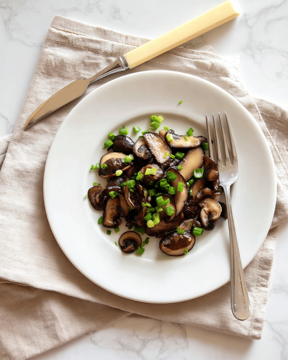 The image shows a white plate with a single layer of cooked dark brown and light brown shiitake mushroom slices spread in the center. Small, bright green chopped green onions are scattered evenly over the mushrooms. The plate sits on a beige cloth napkin, and a silver fork with a pale yellow handle is placed above the plate on the napkin. The entire setting is on a white marbled textured surface. Photo taken with an iphone --ar 4:5 --v 7