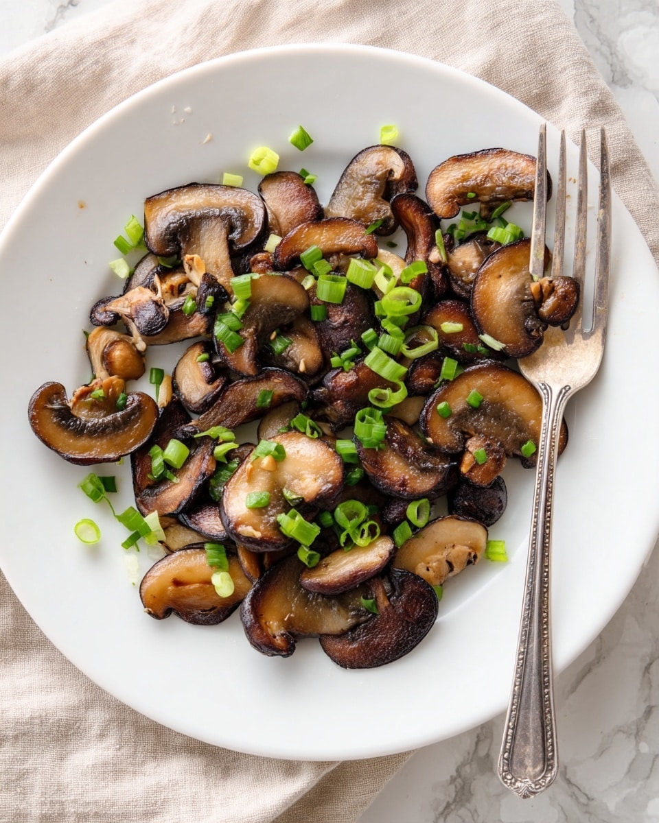 A white plate holds a single layer of cooked mushroom slices, rich brown with dark edges and a soft, slightly glossy texture. Scattered over the mushrooms are small bright green pieces of chopped green onions adding freshness and contrast. A silver fork rests on the right side of the plate, slightly lifting some mushrooms. The plate is set on a white marbled surface with a beige cloth partly visible in the background. Photo taken with an iphone --ar 4:5 --v 7