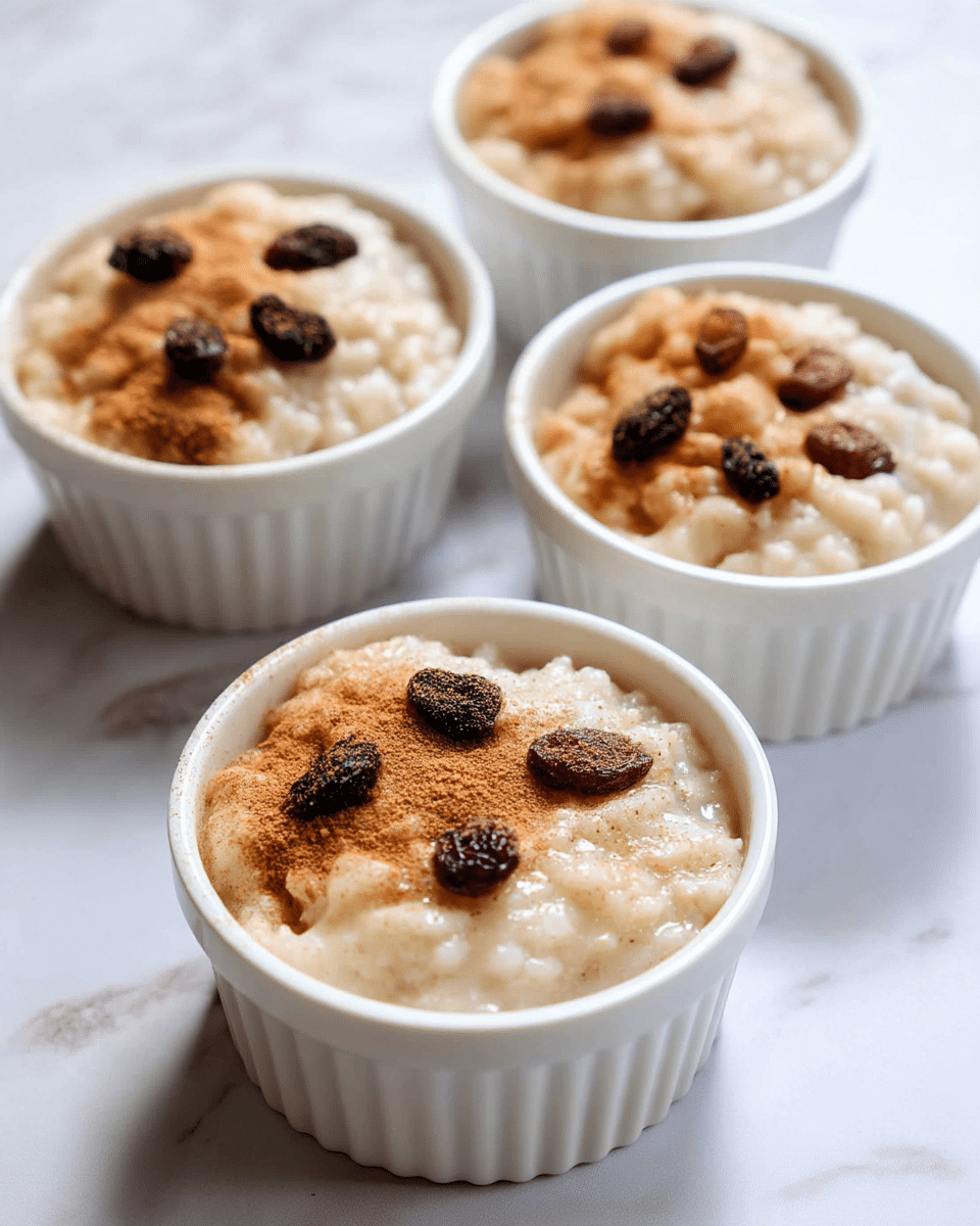 The image shows four white ramekins filled with creamy rice pudding. Each ramekin has a thick, grainy texture of off-white rice pudding with a smooth, slightly shiny surface. On top, there is a light dusting of brown cinnamon powder. Scattered over the cinnamon, there are dark brown raisins, around three to four in each ramekin, placed unevenly near the center. The ramekins are set on a white marbled surface that adds subtle light gray pattern details. The photo is close-up, showing the creamy texture and gentle color contrast between the pudding, cinnamon, and raisins. photo taken with an iphone --ar 4:5 --v 7