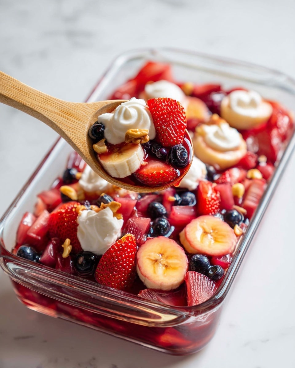 This dish is a colorful fruit salad in a clear glass rectangular container placed on a white marbled surface. The salad has multiple layers of red and purple mixed fruits including sliced strawberries, banana rounds, blueberries, and diced pieces that look like rhubarb or similar fruit, all coated in a glossy, syrupy red liquid. On top, there are small dollops of white whipped cream and small pieces of crushed nuts scattered around. A light wooden spoon is scooping some of the salad from the bottom right corner of the container. photo taken with an iphone --ar 4:5 --v 7