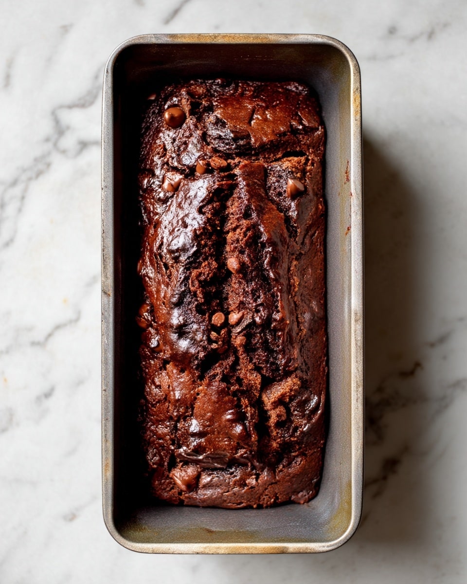 A dark chocolate loaf cake is shown in a rectangular metal baking pan on a white marbled surface. The cake has a rich brown color with a slightly cracked top, revealing moist textures beneath. Small chocolate chips are visible throughout the dense cake, adding texture and contrast to the shiny, somewhat rough surface. The pan has worn edges, showing light discoloration from baking use. photo taken with an iphone --ar 4:5 --v 7
