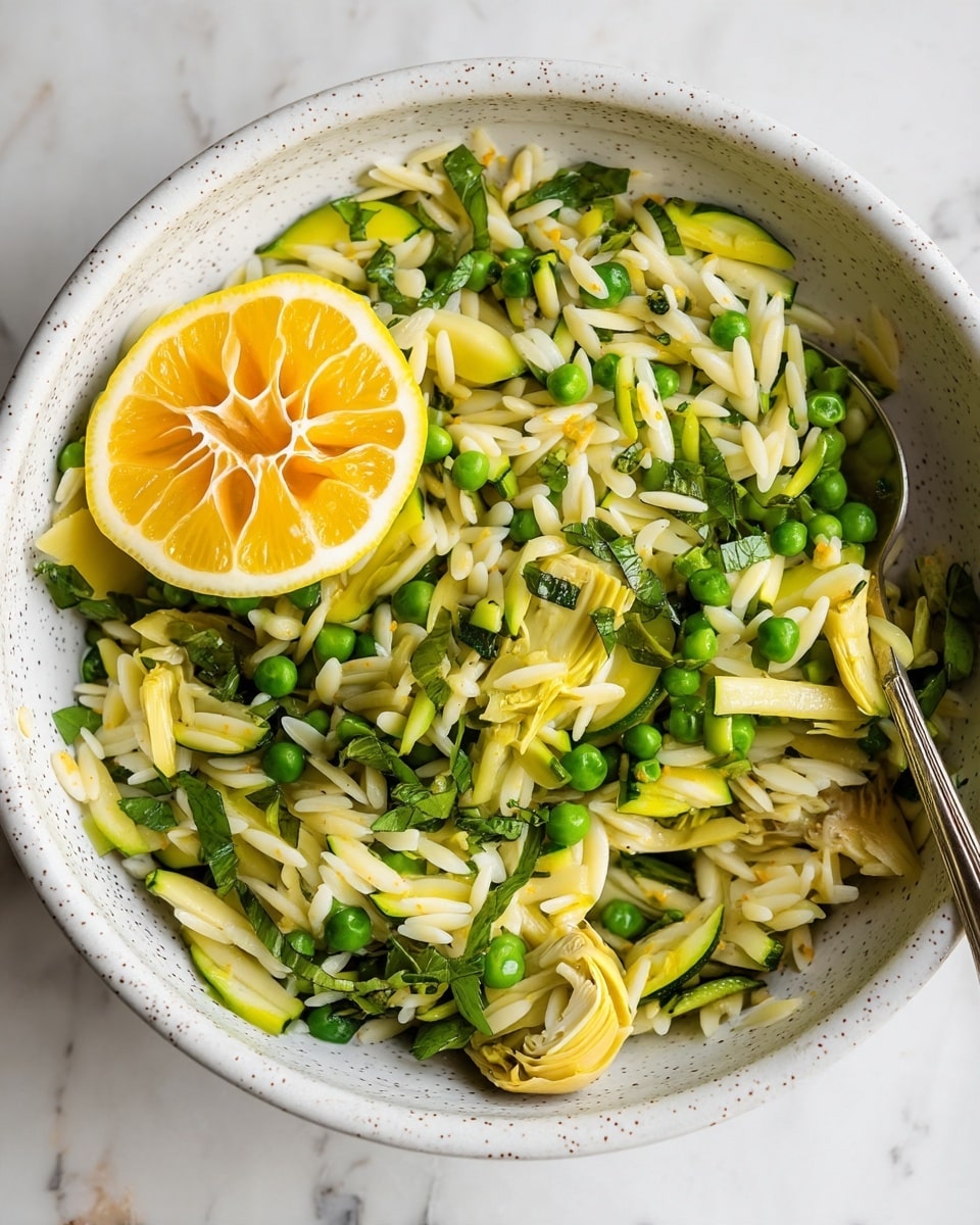 A white speckled bowl holds a fresh orzo pasta salad layered with small light yellow artichoke hearts, bright green peas, thin strips of zucchini, and chopped leafy green herbs scattered throughout. Near the top left of the bowl, a bright yellow lemon half rests on the orzo, adding a burst of color. The textures range from the smooth, glossy orzo grains to the soft artichoke pieces and fresh vegetable bits. A silver spoon peeks out from the right side, partially buried under the salad. The bowl is set on a white marbled surface. photo taken with an iphone --ar 4:5 --v 7